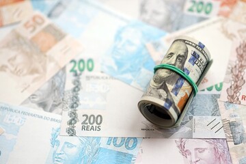 Roll of US dollar bills lies on big amount of Brazilian reais banknotes close up