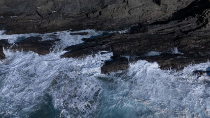 Rocky coastline with powerful waves crashing against rugged cliffs under a clear sky, showcasing the contrast between deep blue ocean and barren land.