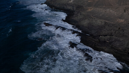 Rocky coastline with powerful waves crashing against rugged cliffs under a clear sky, showcasing the contrast between deep blue ocean and barren land.
