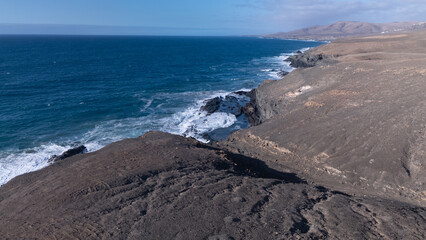Rocky coastline with powerful waves crashing against rugged cliffs under a clear sky, showcasing the contrast between deep blue ocean and barren land.