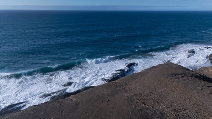 Rocky coastline with powerful waves crashing against rugged cliffs under a clear sky, showcasing the contrast between deep blue ocean and barren land.