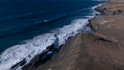 Rocky coastline with powerful waves crashing against rugged cliffs under a clear sky, showcasing the contrast between deep blue ocean and barren land.