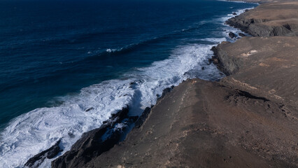 Rocky coastline with powerful waves crashing against rugged cliffs under a clear sky, showcasing the contrast between deep blue ocean and barren land.