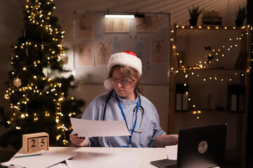 Overworked nurse or doctor Reading Documents, doing paperwork while working using laptop computer on night shift in the clinic on christmas eve. Healthcare and medicine