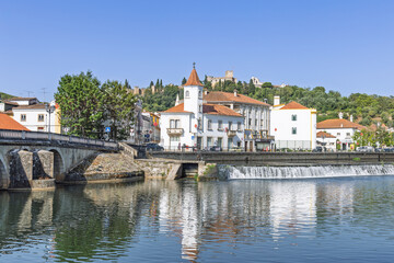 The serene Nabão River flows past a charming Portuguese town, its buildings reflecting in the calm water. The majestic Tomar Castle sits on the hill above, a historic centerpiece.