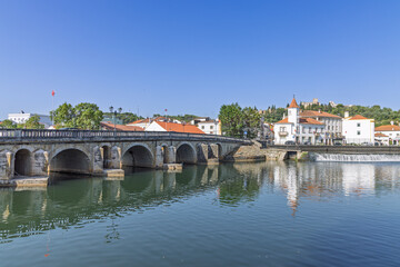 Obraz premium An ancient stone bridge with multiple arches spans the Nabão River in Tomar. The town's historic buildings and the castle on the hill are reflected in the calm water.
