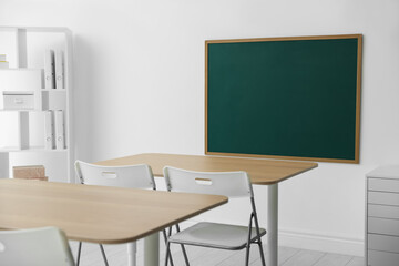 Blank green chalkboard, desks, chairs and shelving unit in classroom