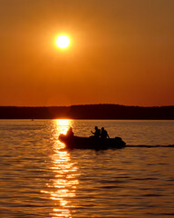 Three people cruise across a tranquil lake in a small motorboat as the sun sets, creating a warm orange glow and reflections on the water's surface