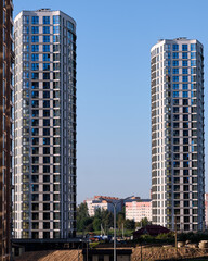 Obraz premium Twin apartment buildings, with repeating window patterns and light gray exteriors, rise above the city landscape under a bright, cloudless blue sky