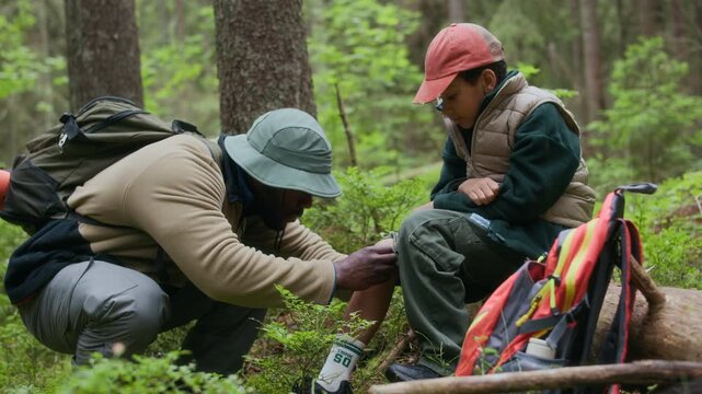 African American father putting bandage on his son scraped knee while hiking in woods, providing care and support during their outdoor adventure
