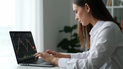 Woman Analyzing Stock Market Data on Laptop - A young woman in a white shirt sits at a desk, intently reviewing candlestick charts on her laptop. - Powered by Adobe