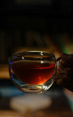 Hand holding clear glass cup of dark tea with lights in background