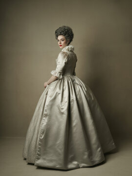 Elegant young woman from the 1700s Rococo era in silver satin pannier gown with lace-trimmed sleeves and powdered coiffure, back three-quarter view portrait