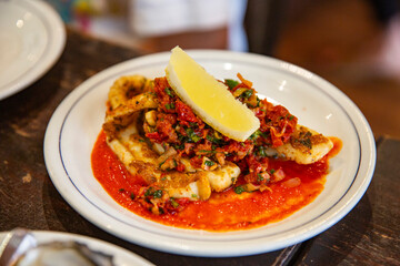 Grilled squid served with red pepper sauce and Mediterranean vegetable garnish, topped with fresh lemon wedge on a white plate in restaurant setting