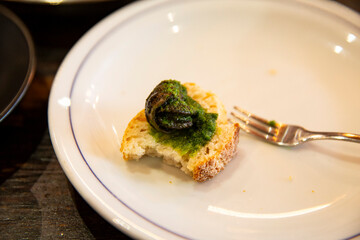 Bite of escargot placed on crusty bread with green herb butter sauce served on a clean white plate with fork in casual French dining setting