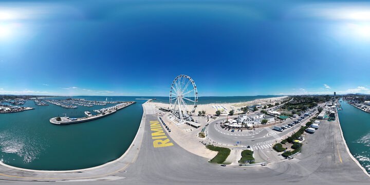 Panoramic view of a vibrant coastal amusement area featuring a large Ferris wheel, marina, sandy beach, and clear blue skies, capturing the essence of summer leisure and fun