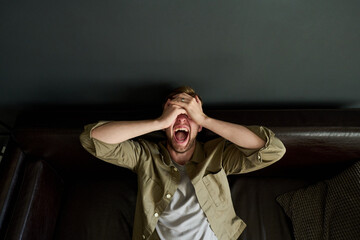 Caucasian young adult man sitting on couch covering face with hands and screaming, expressing...