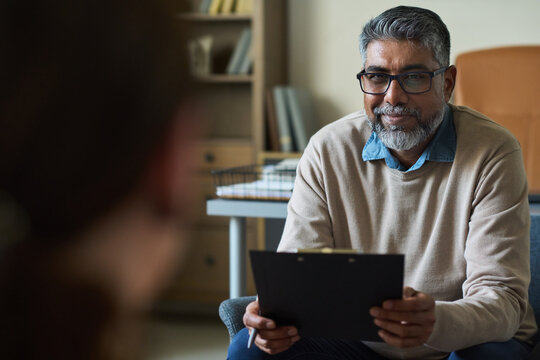 Middle aged South Asian man with gray beard holding clipboard, sitting and attentively listening to unseen in office setting, wearing glasses and smiling gently - Powered by Adobe