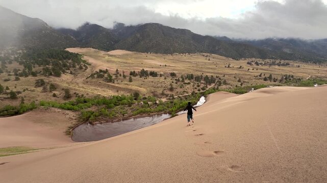 Person running down the Great Sand Dunes National Park Sangre de Cristo range Rocky Mountains spring summer raining cloudy Colorado unique magical large amounts of sand dune hills windy landscape pan