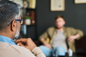 Middle aged South Asian man with gray hair and glasses listening attentively to young Caucasian man during counseling session in office setting, both seated and engaged in conversation