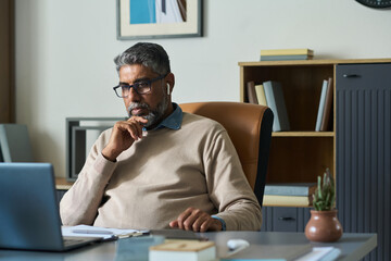 Middle aged South Asian man sitting at desk working on laptop, wearing eyeglasses and wireless earphones, resting chin on hand, appearing focused in modern office setting
