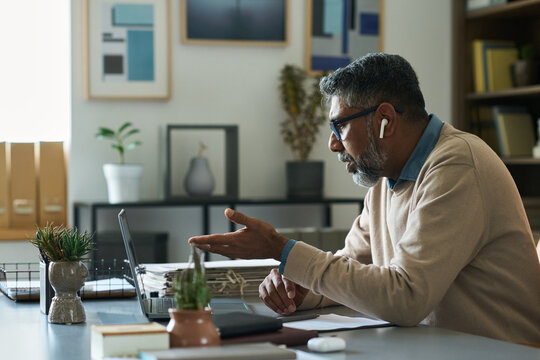 Middle aged Indian man with gray beard sitting at desk gesturing while having video call on laptop, wearing eyeglasses and wireless earbuds, modern office background visible