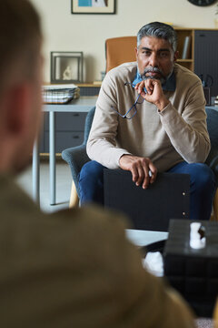 Middle aged South Asian man sitting in office holding eyeglasses and clipboard listening attentively to unseen during counseling or consultation session