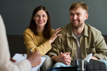 Caucasian young adult woman and Caucasian young adult man sitting together holding hands smiling while attending counseling session with professional writing on clipboard
