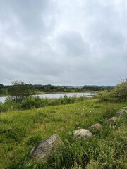small lake surrounded by green grass, bushes and trees in a flat landscape with cloudy sky