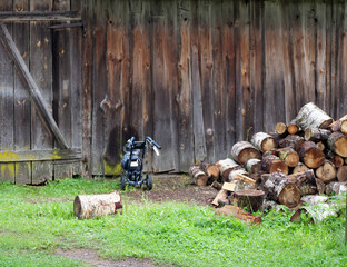Firewood Chopper or Log Splitter in a Rural Backyard - log splitter is sitting on the grass next to a rustic wooden wall and a pile of freshly cut logs.