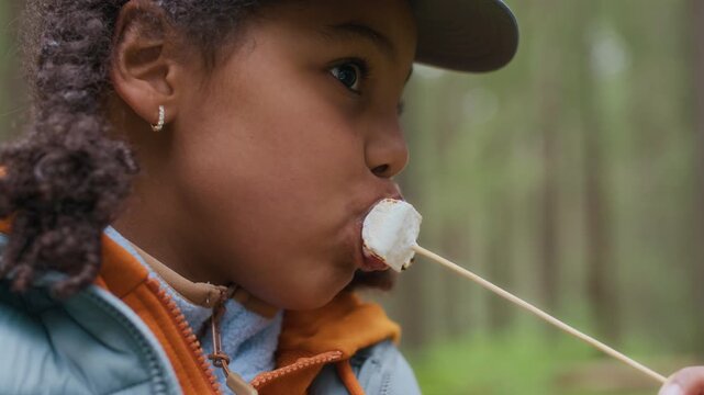 Medium closeup of young African American girl enjoying eating roasted marshmallow while camping in forest, savoring sweet outdoor treat