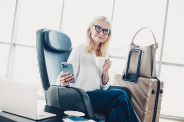 Confident woman at airport terminal, holding a smartphone and coffee, ready for her next travel, sitting near suitcase