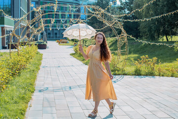 A woman in a light yellow dress with an openwork umbrella in her hand. Summer walk in the recreation area. Traveling light. A young woman walks on a hot day with an  The umbrella is raised above her h