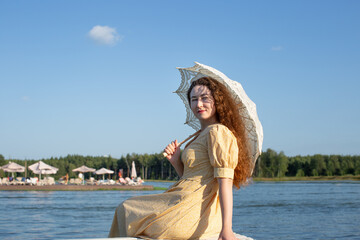 A young woman is sitting in a half-turn with an umbrella in her hand against the background of a summer landscape with a lake. A woman in a light yellow dress with a summer umbrella