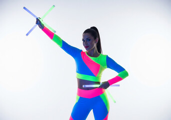 Woman in bright neon athletic wear poses with illuminated batons against a white background, one arm raised.