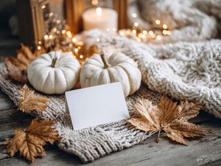 Fototapeta premium Cozy autumn scene with white pumpkins, dried leaves, and blank card on knitted blanket, surrounded by warm candlelight and fairy lights