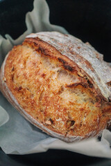 A rustic, golden-brown sourdough loaf sits on parchment paper, showcasing its crust and airy interior. It's beautifully lit against a dark background.
