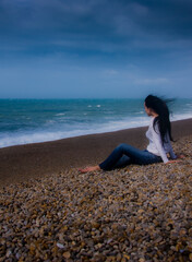 A woman sits on a pebble beach, looking out at the ocean.