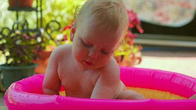 A child is sitting in a bright pink inflatable pool, holding a blue toy in his hands. A stage surrounded by lush green plants. Close-up of a boy sitting in a backyard pool