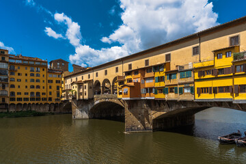 Fototapeta premium Picturesque view of a historical bridge with colorful buildings spanning a river under a sunny sky with scattered clouds.