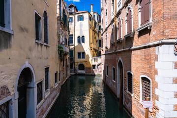 Venetian canal scene with buildings, featuring a narrow waterway reflecting sunlight and architectural details on aged facades.