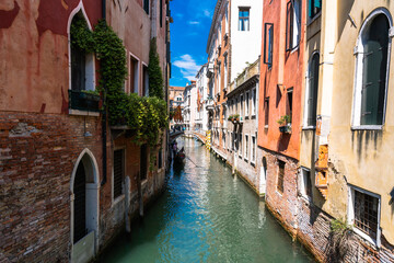 Venetian canal vista: Gondola glides through tranquil waters lined by aged buildings under a bright blue sky. Timeless European charm.