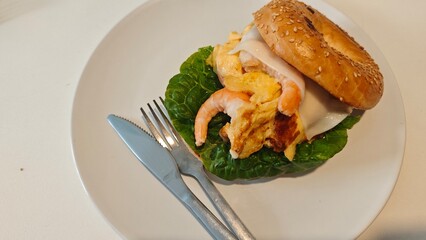 Bagel sandwich with scrambled eggs, shrimp, cheese, and lettuce served on a white plate
