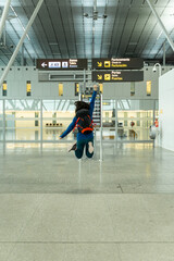 Young woman jumping at the airport, happy to be traveling. She is carrying a large backpack and wearing a blue sweatshirt.