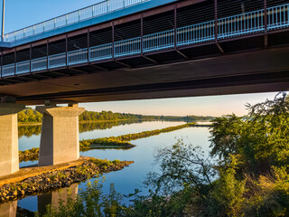 Under the bridge shot highlights its steel structure and concrete pillars, with the Vistula river and distant shoreline visible.