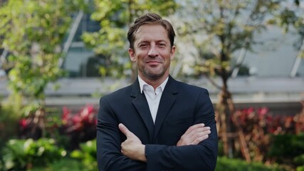 Portrait of happy businessman in suit crosses arms standing in urban park. Mature man entrepreneur smiles conveying success and professionalism in downtown district slow motion