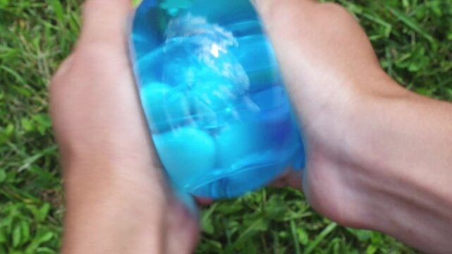 Press the hands tightly and twist the translucent blue slime ball above green grass, the elastic surface is deformed noticeably under the daylight. Antisters. ASMR.