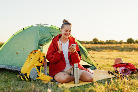 A cheerful young woman claps and smiles during a video call on her smartphone while camping by her tent.