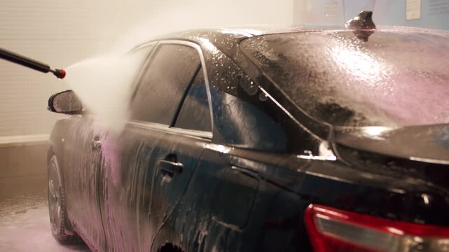 Working with a Karcher to wash a black car in a garage. A jet of water under pressure washes away pink foam and dirt from the car and glass. Self-service car wash as a profitable business