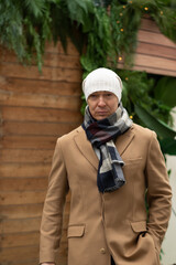 Naklejka premium Winter portrait of caucasian man wearing white beanie and cozy scarf, with blurred Christmas festive background. Cold winter weather. Outdoors. Xmas holiday.